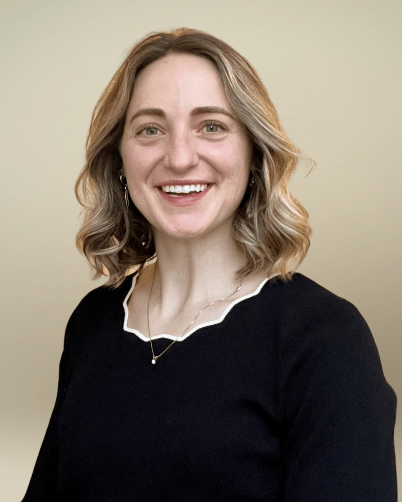 Portrait of a smiling woman with shoulder-length wavy blonde hair wearing a black top with a scalloped neckline, photographed against a neutral background.